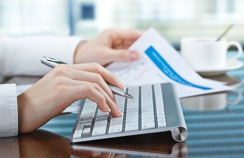 Woman's hand holding pen and report while typing on keyboard